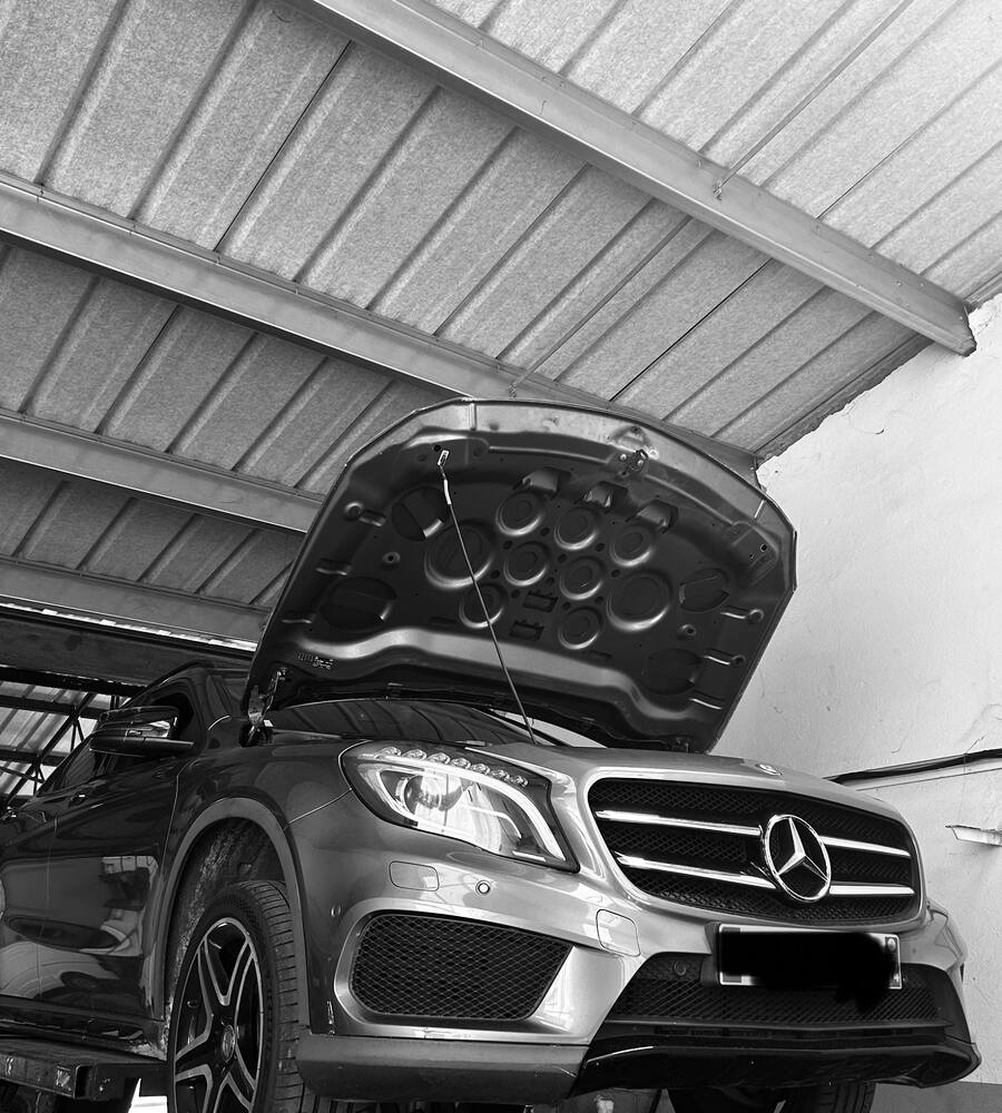 Une Mercedes grise levée sur le pont, capot ouvert, sous la toiture en tôle de l’atelier du Garage KNR